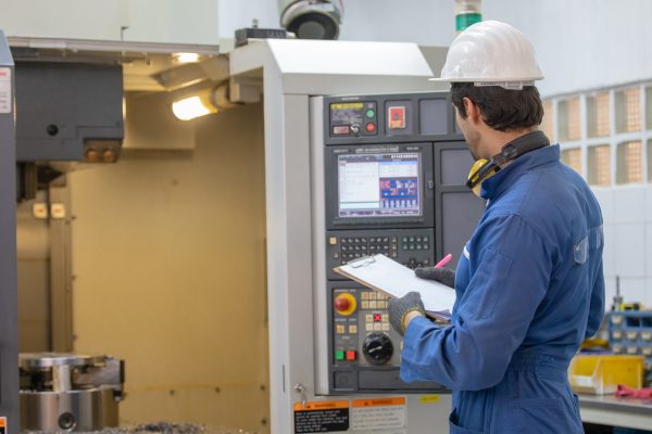 an engineer wearing a white hard hat in a smart factory standing in front of a control panel inspecting the machine 