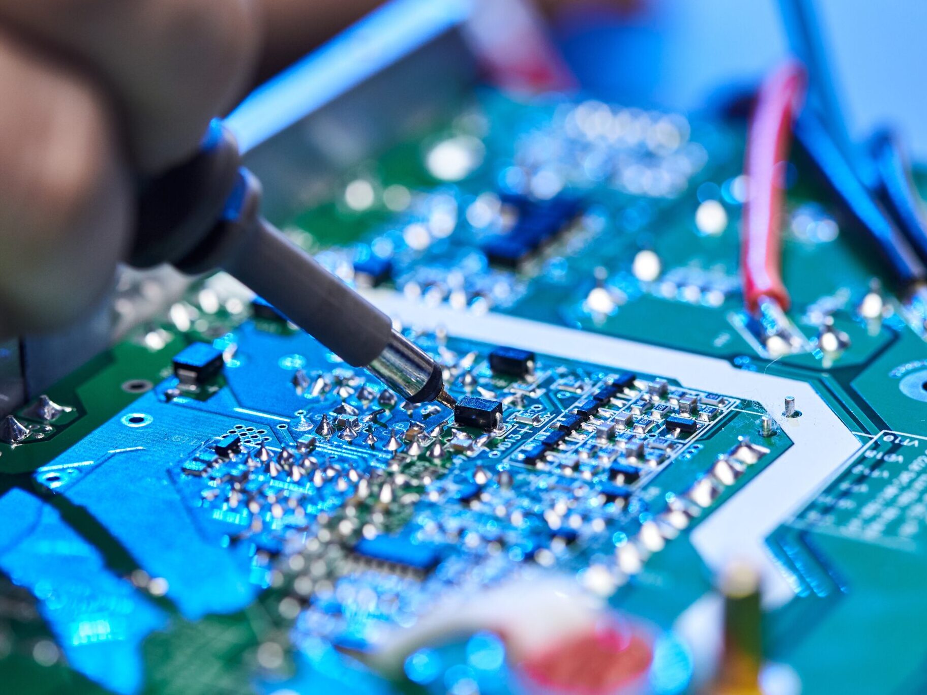Close-up of a technician using a probe on a brightly lit blue and green printed circuit board (PCB), representing the use of AI vision for quality inspection, micro-component verification, and defect analysis in the electronics industry.