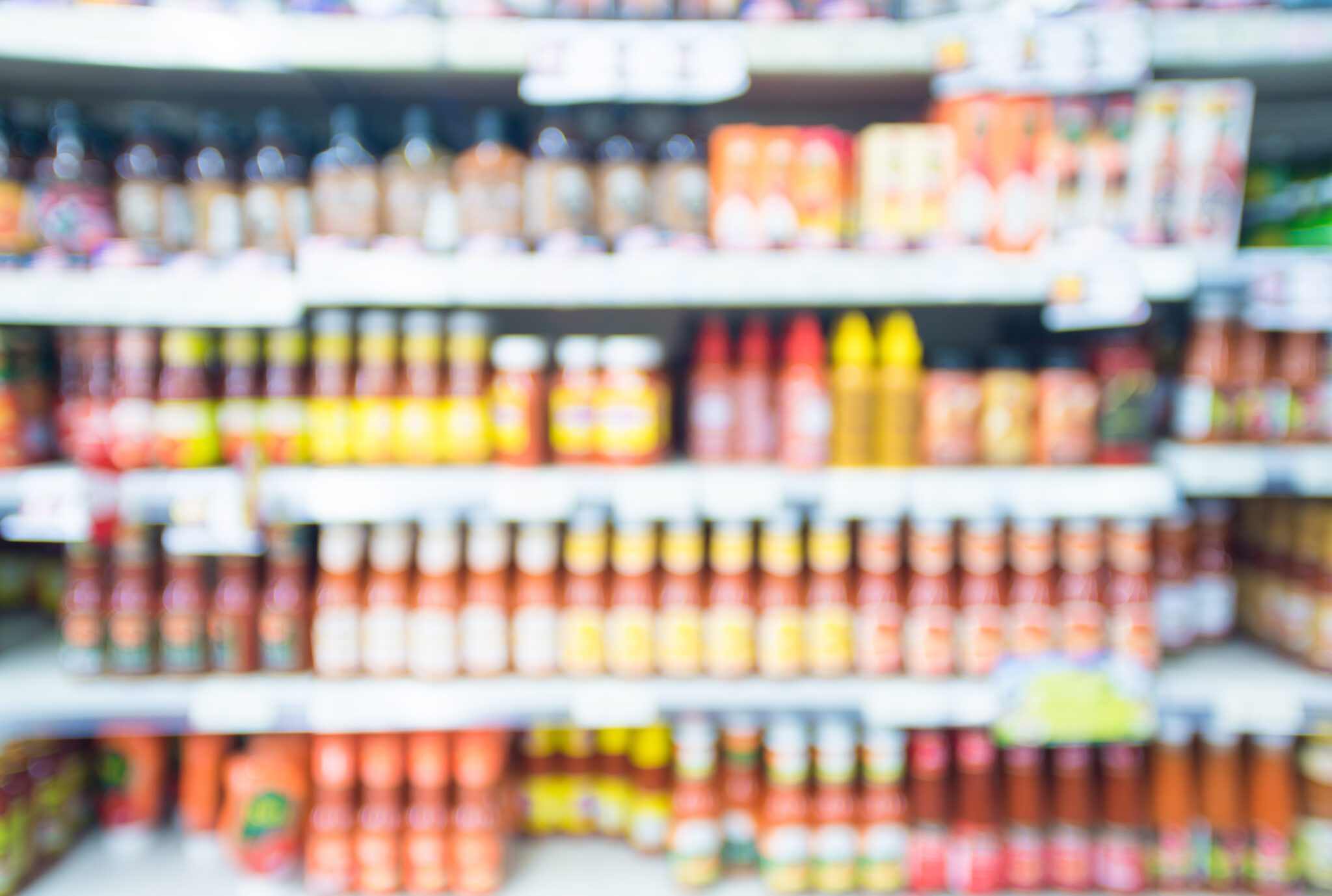 Blurred image of fully stocked supermarket shelves with various packaged goods and sauces, representing the use of AI vision in the retail sector for inventory management, on-shelf stock detection, and automated self-checkout.