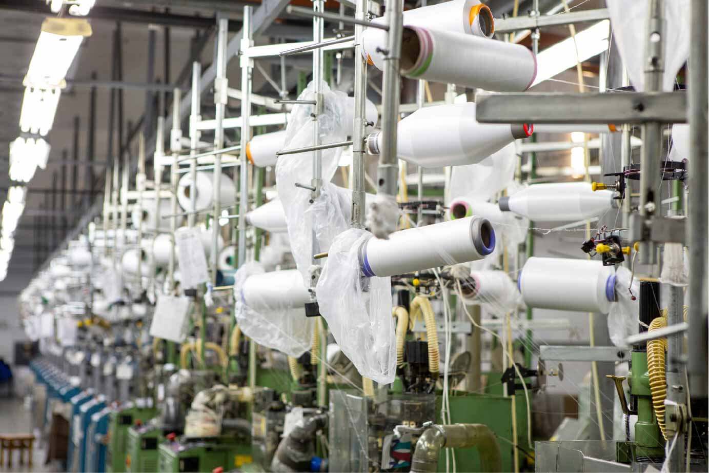 Brightly lit factory floor showing rows of automated textile machinery with numerous spools of white yarn or thread, illustrating the application of AI vision for process monitoring, quality control, and defect detection in the textiles and footwear manufacturing industry.