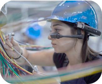 Human-AI Collaboration via Augmented Intelligence for Frontline Workers A female frontline worker wearing a blue hard hat and a wearable AR glasses headset while inspecting complex electrical wiring, demonstrating augmented intelligence and human-AI collaboration in an industrial environment.
