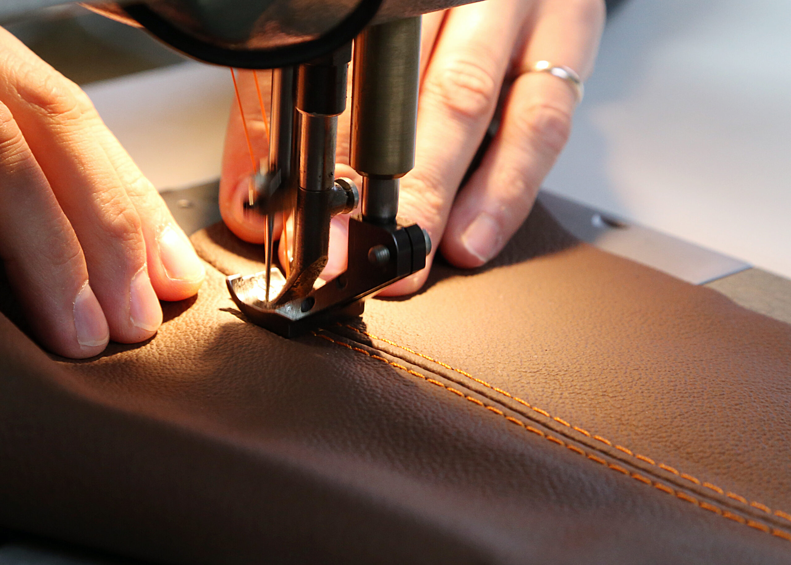 A close-up view of a craftsman's hands carefully guiding a piece of brown leather with distinct orange stitching under the needle of an industrial sewing machine. This emphasizes the role of AI vision in the luxury goods industry for ensuring quality, defect detection in high-value materials (like leather), and handling precious items.